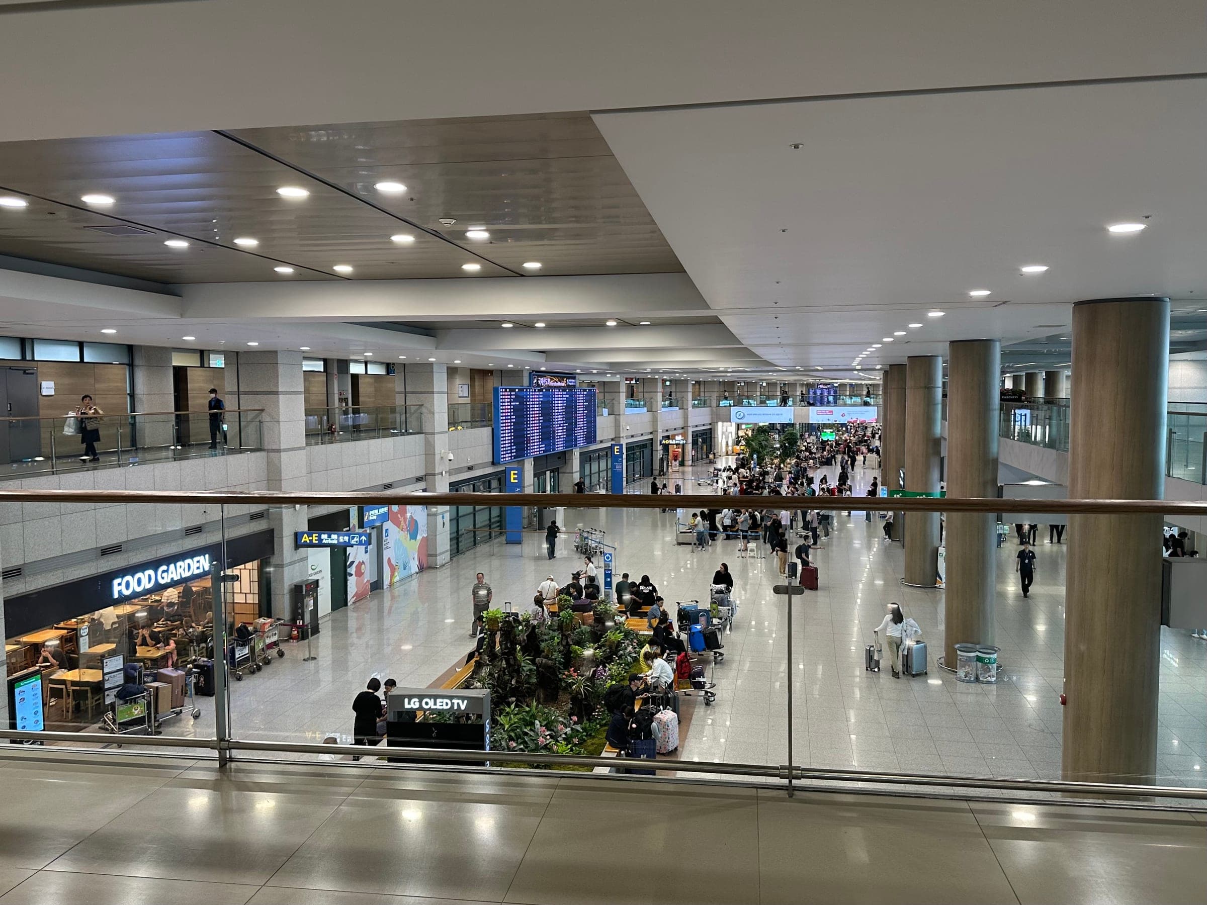 The arrival floor of Incheon Airport Terminal 1 seen from the level above, with a flight board and travellers with luggage