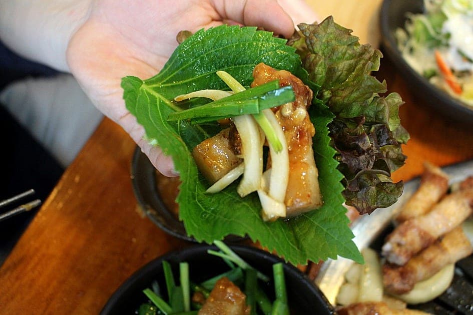 A hand holding a perilla leaf and lettuce wrap filled with grilled pork belly, onion and spring onion