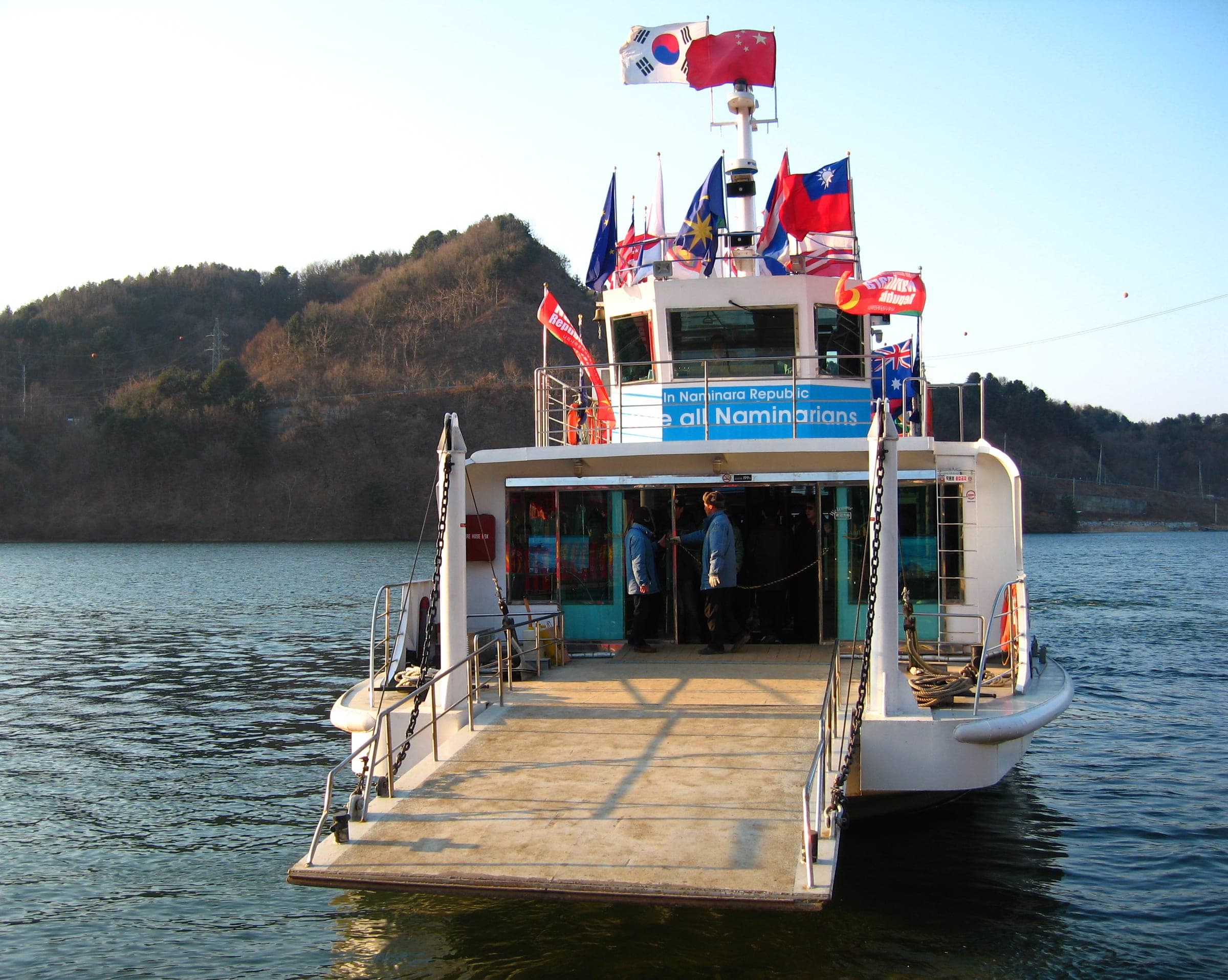 A white ferry decked with national flags, its boarding ramp lowered, on the Bukhan River near Nami Island