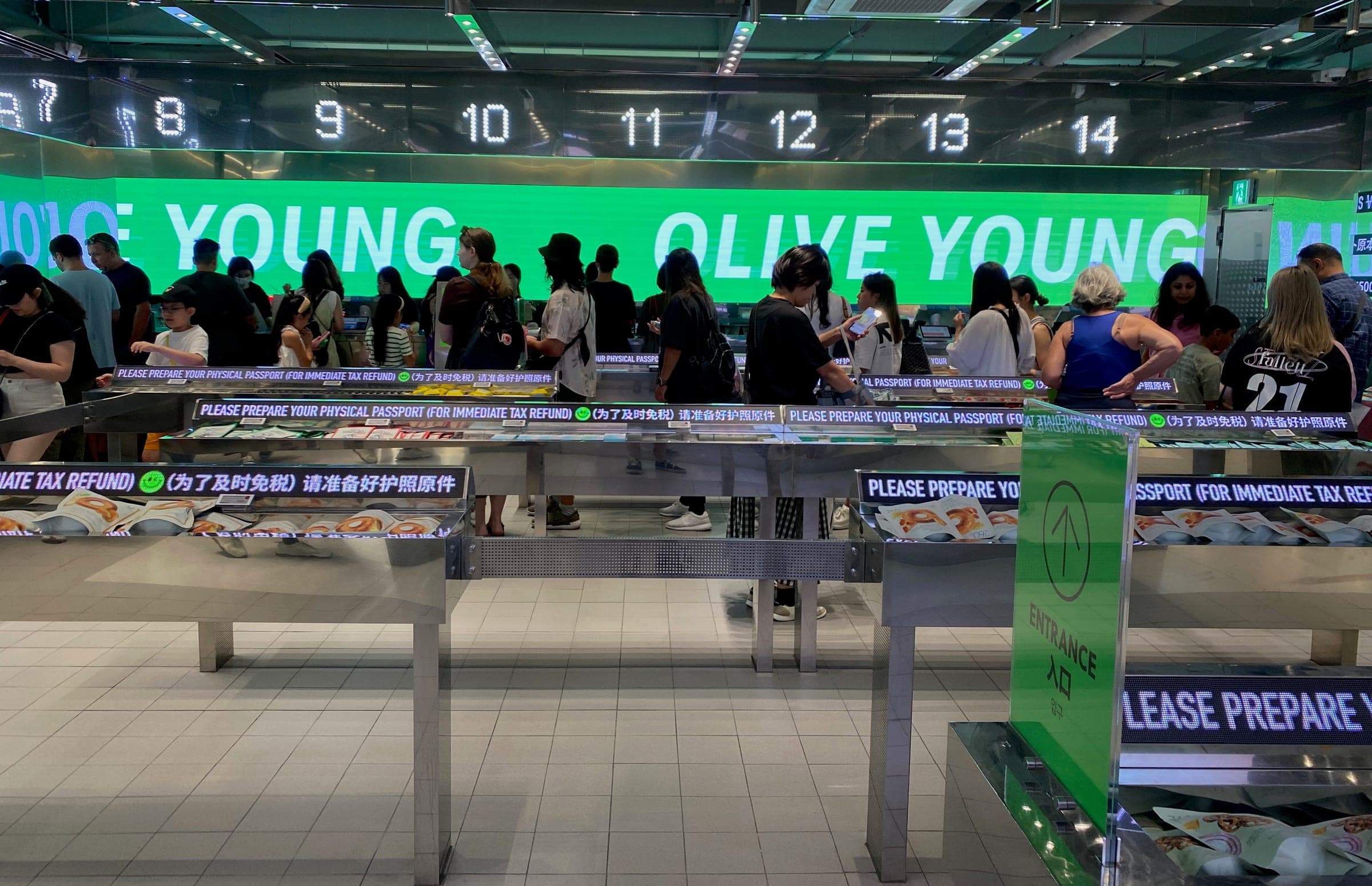 Shoppers queueing at numbered checkout counters in an Olive Young store in Myeongdong, with signs asking customers to prepare a physical passport for immediate tax refund